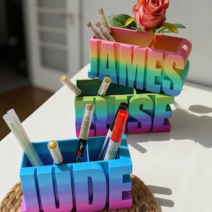 Pu&ograve; includere: Tre organizer da scrivania personalizzati con un design a gradiente arcobaleno. Gli organizer scrivono "JAMES", "ELISE" e "JUDE" in grandi lettere. Ogni organizer contiene penne e pennarelli. Una rosa rossa &egrave; in cima all'organizer "JAMES".