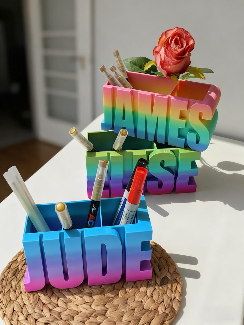 Pu&ograve; includere: Tre organizer da scrivania personalizzati con un design a gradiente arcobaleno. Gli organizer scrivono "JAMES", "ELISE" e "JUDE" in grandi lettere. Ogni organizer contiene penne e pennarelli. Una rosa rossa &egrave; in cima all'organizer "JAMES".