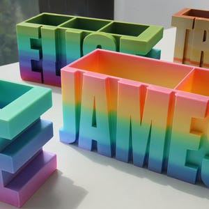 Pu&ograve; includere: Organizzatori da scrivania colorati che compongono nomi. Gli organizzatori sono a forma di lettere a blocchi, ogni lettera di un colore diverso dell'arcobaleno. I nomi sono "JAMES", "ELIJAH" e "TRISTAN". Hanno scomparti per penne, matite e altri materiali.