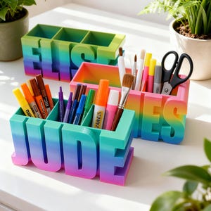 May include: Three vibrant desk tidies, spelling "JUDE", "JAMES", and "ELIS". The organisers feature a gradient of colours, from purple to green. Each tidy holds pens, markers, brushes, and scissors. A creative and functional desk accessory, ideal for stationery.