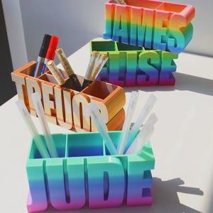 Pu&ograve; includere: Tre portapenne personalizzati in una sfumatura arcobaleno. I nomi "JAMES", "ELISE", "TREVOR" e "JUDE" sono visualizzati in grandi lettere. Ogni supporto ha scomparti per penne e pennarelli.