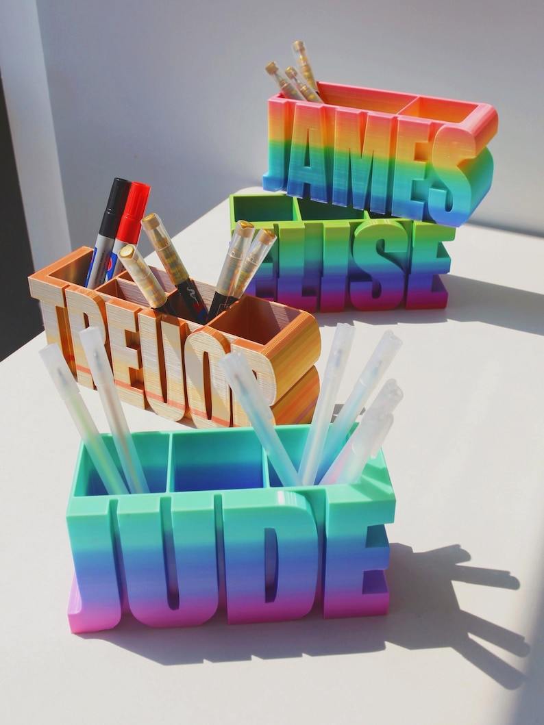 Pu&ograve; includere: Tre portapenne personalizzati in una sfumatura arcobaleno. I nomi "JAMES", "ELISE", "TREVOR" e "JUDE" sono visualizzati in grandi lettere. Ogni supporto ha scomparti per penne e pennarelli.