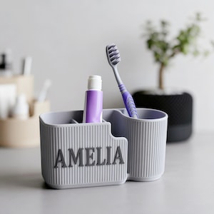Könnte beinhalten: Ein grauer Zahnbürstenhalter mit dem Namen "AMELIA" in dunkelgrauen Buchstaben. Der Halter enthält eine Tube lila Zahnpasta und eine lila und graue Zahnbürste. Der Halter hat ein geripptes Design.