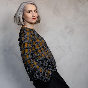 Puede incluir: Una mujer con un suéter calado negro y gris sobre una blusa mostaza y una falda negra. El suéter tiene un patrón de rejilla. La mujer tiene el pelo corto gris y lápiz labial rojo. El fondo es una pared gris.