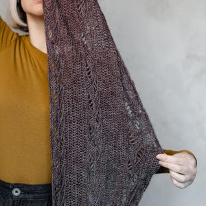 Puede incluir: Un chal de tejido abierto gris oscuro con un delicado patrón de encaje. El chal se sostiene, revelando su intrincado diseño y textura. El fondo es gris neutro, resaltando el color y los detalles del chal. El chal parece hecho a mano.