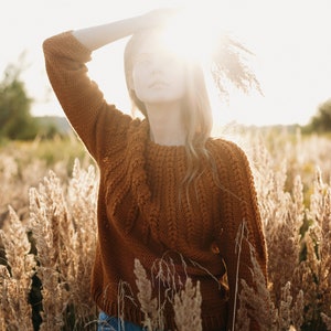 May include: A woman wearing a brown knitted sweater and blue jeans stands in a field of tall grass. The sun shines brightly behind her, creating a halo effect around her head.