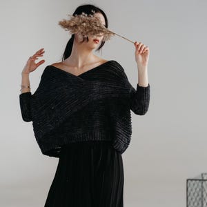 Könnte beinhalten: Ein dunkelgrauer Off-Shoulder-Pullover mit strukturiertem Rippendesign. Der Pullover wird mit einem langen, schwarzen Faltenrock kombiniert. Das Model hält eine getrocknete Pflanze vor seinem Gesicht.