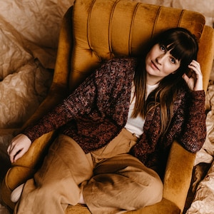 Puede incluir: Una mujer con un cárdigan marrón y morado, pantalones beige y zapatillas blancas se sienta en un sillón amarillo mostaza. El sillón está rodeado de papel marrón arrugado.