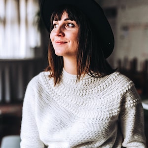 Op de afbeelding: Een vrouw draagt een witte gebreide trui met een zwarte hoed. De trui heeft een gedetailleerd gebreid patroon rond de halslijn.