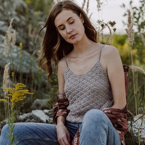 May include: A woman wearing a light brown crochet tank top, blue jeans, and a brown floral patterned shirt. She is sitting on a rocky surface with tall grasses and flowers in the background.