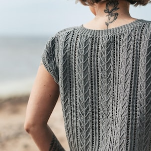 May include: A grey short-sleeved top featuring a vertical knit pattern with repeating leaf-like textures and a round neckline. The garment has a structured ribbed hem and a fitted silhouette, showcasing a detailed stitch design throughout the fabric.