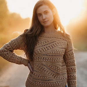 May include: A woman wearing a brown crocheted sweater with a lacy pattern. She is standing in a field with the sun setting in the background.