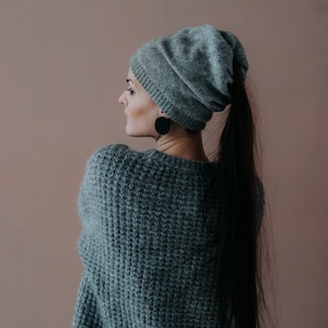 May include: A woman with long dark hair is wearing a grey slouchy beanie and a grey sweater. She is standing in front of a light brown wall.