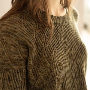 Może przedstawiać: Oliwkowy sweter z dzianiny o teksturowanym, wzorzystym designie. Sweter posiada okrągły dekolt i długie rękawy. Wzór dzianiny tworzy wzór przypominający liście na przodzie i rękawach. Sweter wykonany jest z miękkiego, ciepłego materiału.