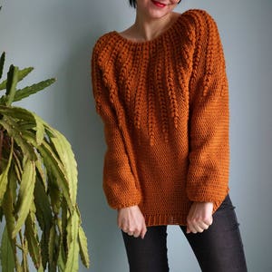 May include: A crocheted sweater in a burnt orange color with a unique stitch pattern. The sweater has a wide neckline and long sleeves.