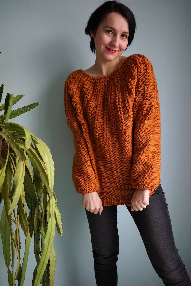 May include: A crocheted sweater in a burnt orange color with a unique stitch pattern. The sweater has a wide neckline and long sleeves.