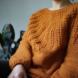 May include: A close-up of a crocheted sweater in a warm orange color. The sweater has a textured stitch pattern and a relaxed fit.