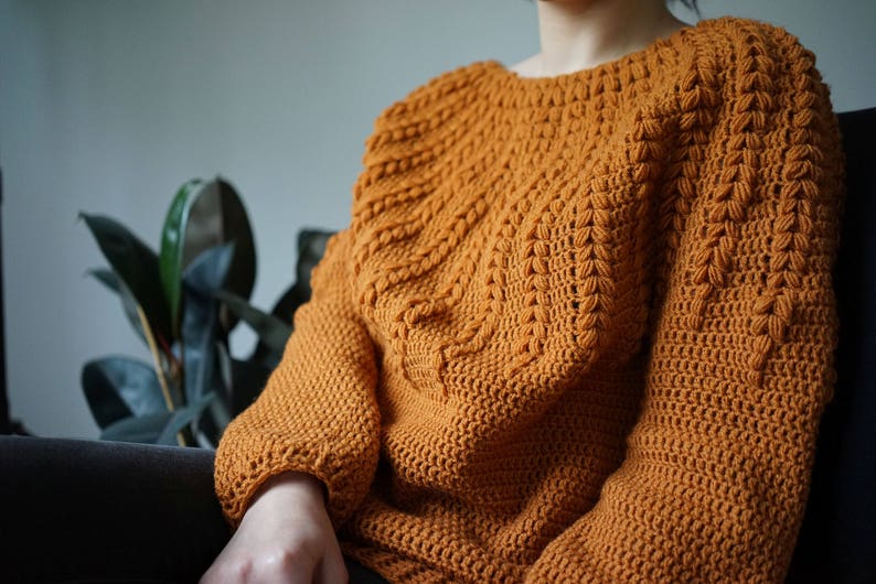 May include: A close-up of a crocheted sweater in a warm orange color. The sweater has a textured stitch pattern and a relaxed fit.