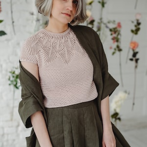 May include: A woman wearing a light pink crocheted top with a delicate leaf pattern and a dark green linen skirt. She is standing in front of a white wall with floral decorations.