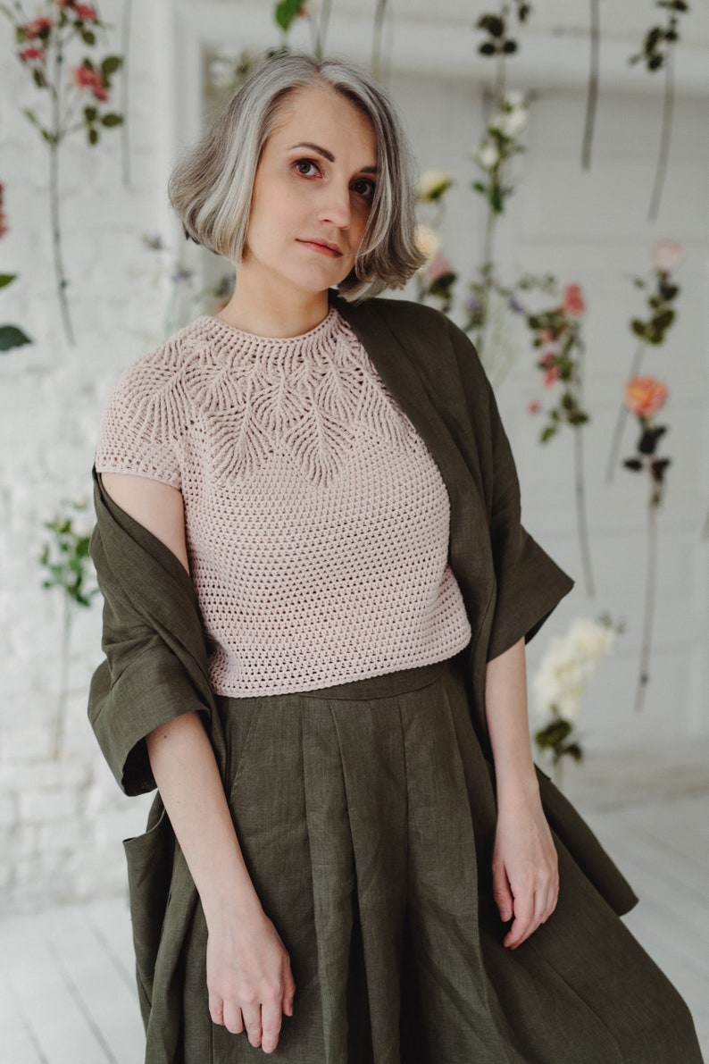May include: A woman wearing a light pink crocheted top with a delicate leaf pattern and a dark green linen skirt. She is standing in front of a white wall with floral decorations.