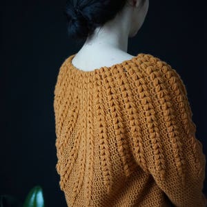 May include: A close-up of a crocheted sweater in a warm mustard yellow color. The sweater has a textured stitch pattern and a loose, comfortable fit.