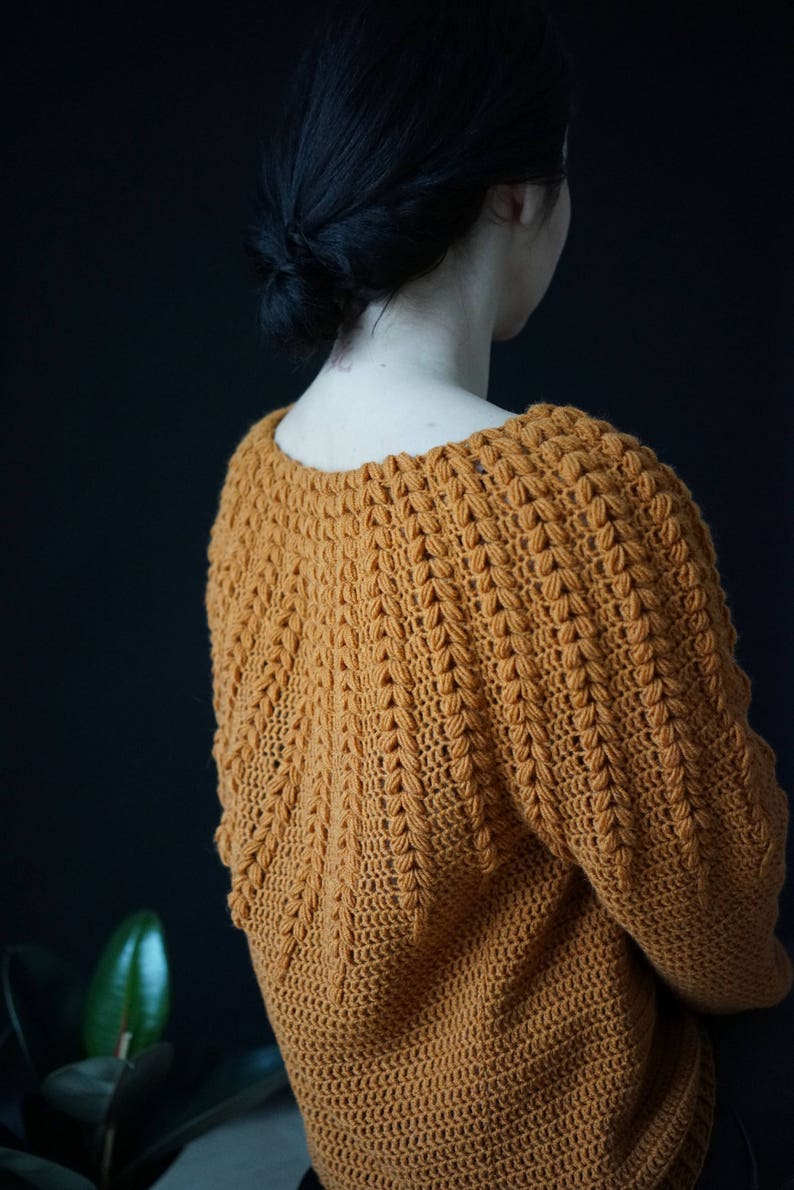 May include: A close-up of a crocheted sweater in a warm mustard yellow color. The sweater has a textured stitch pattern and a loose, comfortable fit.