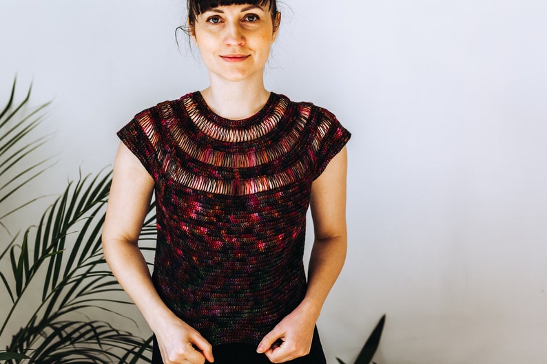 May include: A woman wearing a short-sleeved, crocheted top with a colorful, multi-colored, and textured design. The top has a wide, crocheted neckline.