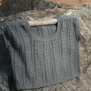 May include: A dark gray sleeveless top featuring a vertical knit pattern with textured ribbing along the scoop neckline and armholes. The garment has a cropped length and a consistent stitch design throughout the front, showcasing a detailed weave.