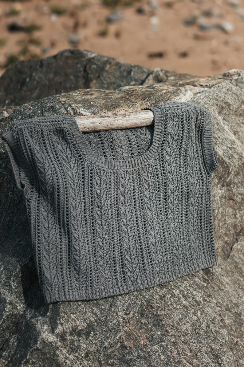 May include: A dark gray sleeveless top featuring a vertical knit pattern with textured ribbing along the scoop neckline and armholes. The garment has a cropped length and a consistent stitch design throughout the front, showcasing a detailed weave.