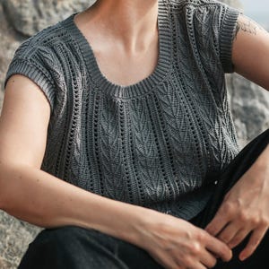 May include: A dark gray short-sleeved knit top featuring a round neckline and a textured pattern of vertical columns. The garment displays a repeating leaf-like stitch design interspersed with vertical eyelet rows, finished with ribbed trim.