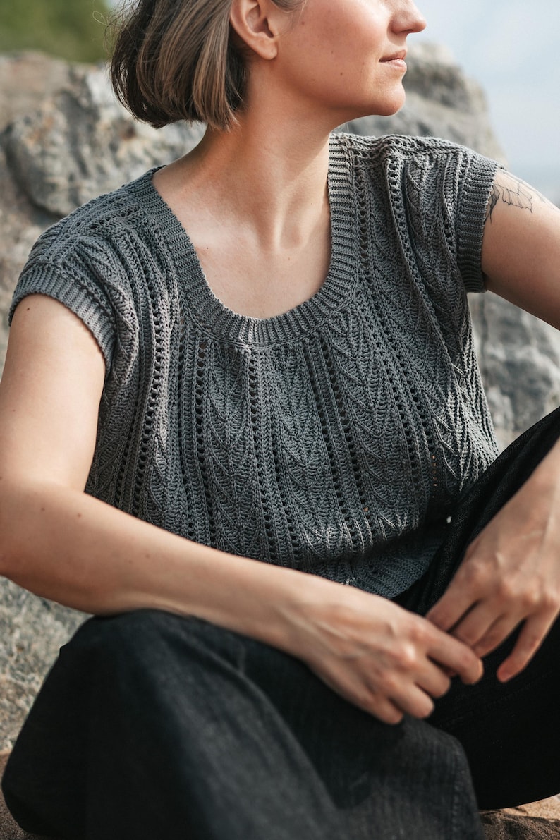 May include: A dark gray short-sleeved knit top featuring a round neckline and a textured pattern of vertical columns. The garment displays a repeating leaf-like stitch design interspersed with vertical eyelet rows, finished with ribbed trim.