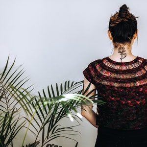 May include: A woman wearing a dark red, black, and green crochet top with a floral design on the back of her neck. The top has a wide, open neckline and short sleeves.