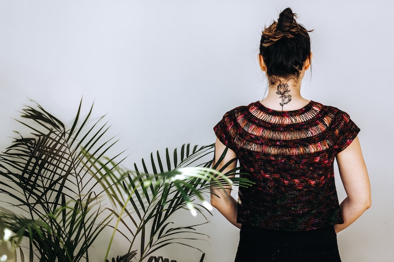 May include: A woman wearing a dark red, black, and green crochet top with a floral design on the back of her neck. The top has a wide, open neckline and short sleeves.