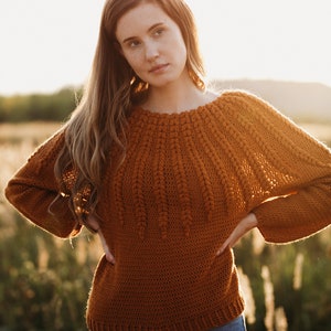May include: A woman wearing a brown crocheted sweater with a unique stitch pattern and blue jeans. She is standing in a field with tall grass.