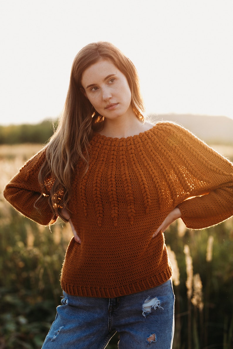May include: A woman wearing a brown crocheted sweater with a unique stitch pattern and blue jeans. She is standing in a field with tall grass.