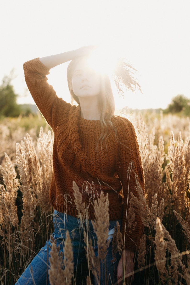 May include: A woman wearing a brown knitted sweater and blue jeans stands in a field of tall grass. The sun shines brightly behind her, creating a halo effect around her head.