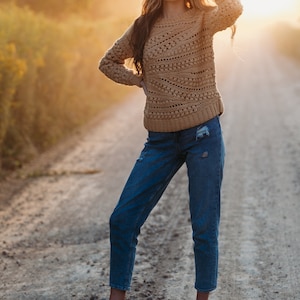 May include: A woman wearing a brown crocheted sweater, blue jeans, and black shoes stands on a dirt road. The sun is setting in the background.