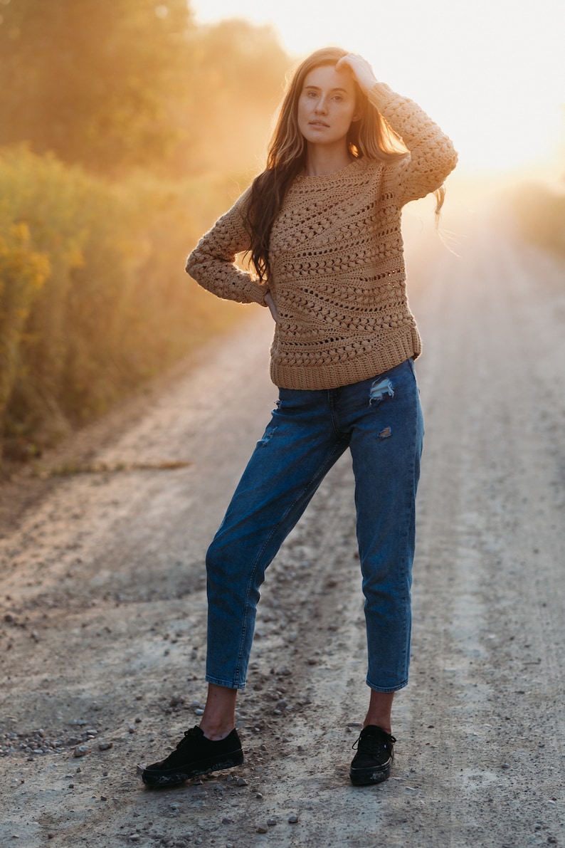 May include: A woman wearing a brown crocheted sweater, blue jeans, and black shoes stands on a dirt road. The sun is setting in the background.