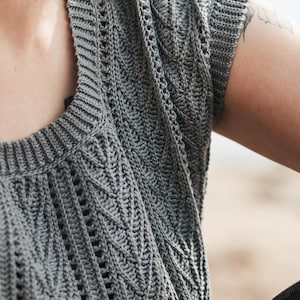 May include: A grey sleeveless top featuring a textured crochet knit pattern with vertical leaf-like motifs and openwork detailing. The garment has a ribbed crew neckline and ribbed armholes, constructed with a consistent stitch throughout the piece.