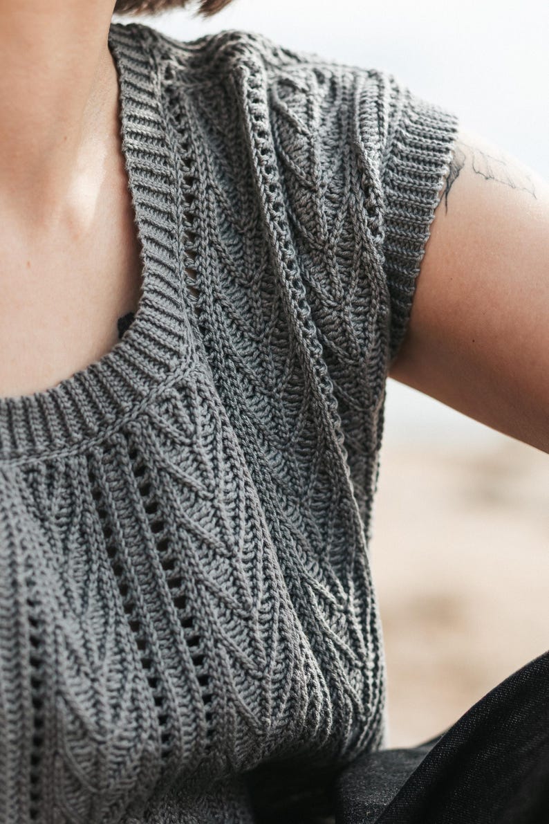 May include: A grey sleeveless top featuring a textured crochet knit pattern with vertical leaf-like motifs and openwork detailing. The garment has a ribbed crew neckline and ribbed armholes, constructed with a consistent stitch throughout the piece.