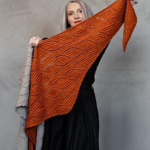 Op de afbeelding: Een oranje gebreide sjaal met een golfpatroon wordt getoond. De sjaal heeft een warme, oranje kleur. Een tweede, grijze sjaal is ook zichtbaar. De sjaals zijn gemaakt van zacht, getextureerd garen.