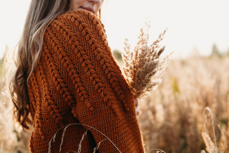 May include: A close-up of a woman wearing a brown crocheted cardigan. The cardigan has a textured pattern and is worn over a white shirt. The woman is holding a bouquet of dried pampas grass.
