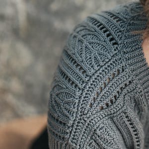 May include: A slate gray short-sleeved top featuring intricate crochet work with a textured leaf-like pattern on the shoulder and upper sleeve. The garment includes vertical ribbed detailing and openwork stitches that create a structured design.
