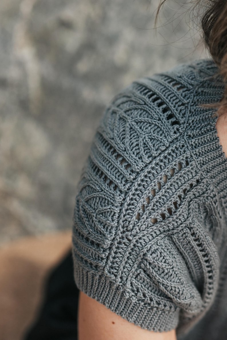 May include: A slate gray short-sleeved top featuring intricate crochet work with a textured leaf-like pattern on the shoulder and upper sleeve. The garment includes vertical ribbed detailing and openwork stitches that create a structured design.