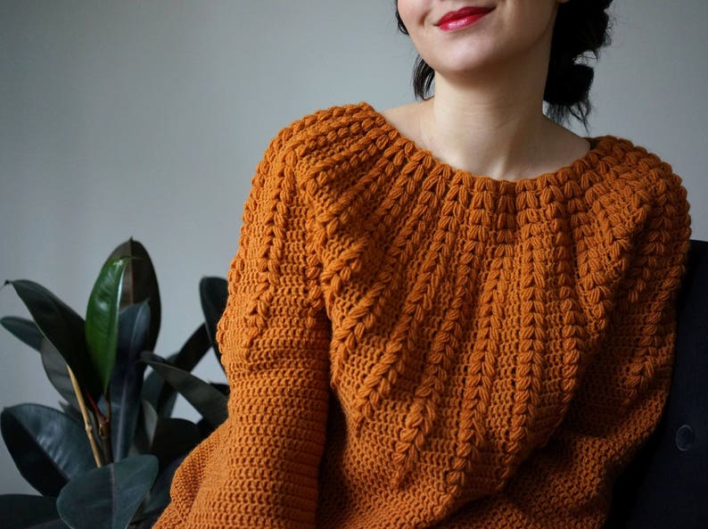May include: A close-up of a crocheted sweater in a warm orange color. The sweater has a unique stitch pattern that creates a textured look. The sweater has a wide neckline and long sleeves.