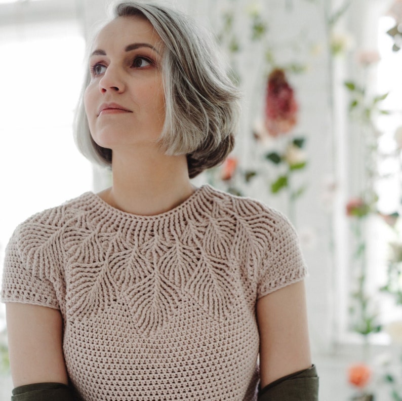 May include: A woman with short, gray hair is wearing a light pink crocheted top with a leaf pattern. The top has short sleeves and a round neckline.