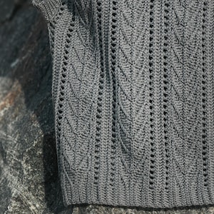 May include: A dark gray sleeveless top featuring a vertical leaf-like crochet pattern with openwork detailing. The garment is constructed with a ribbed hem and a textured knit design that runs throughout the fabric, creating a repeating motif.