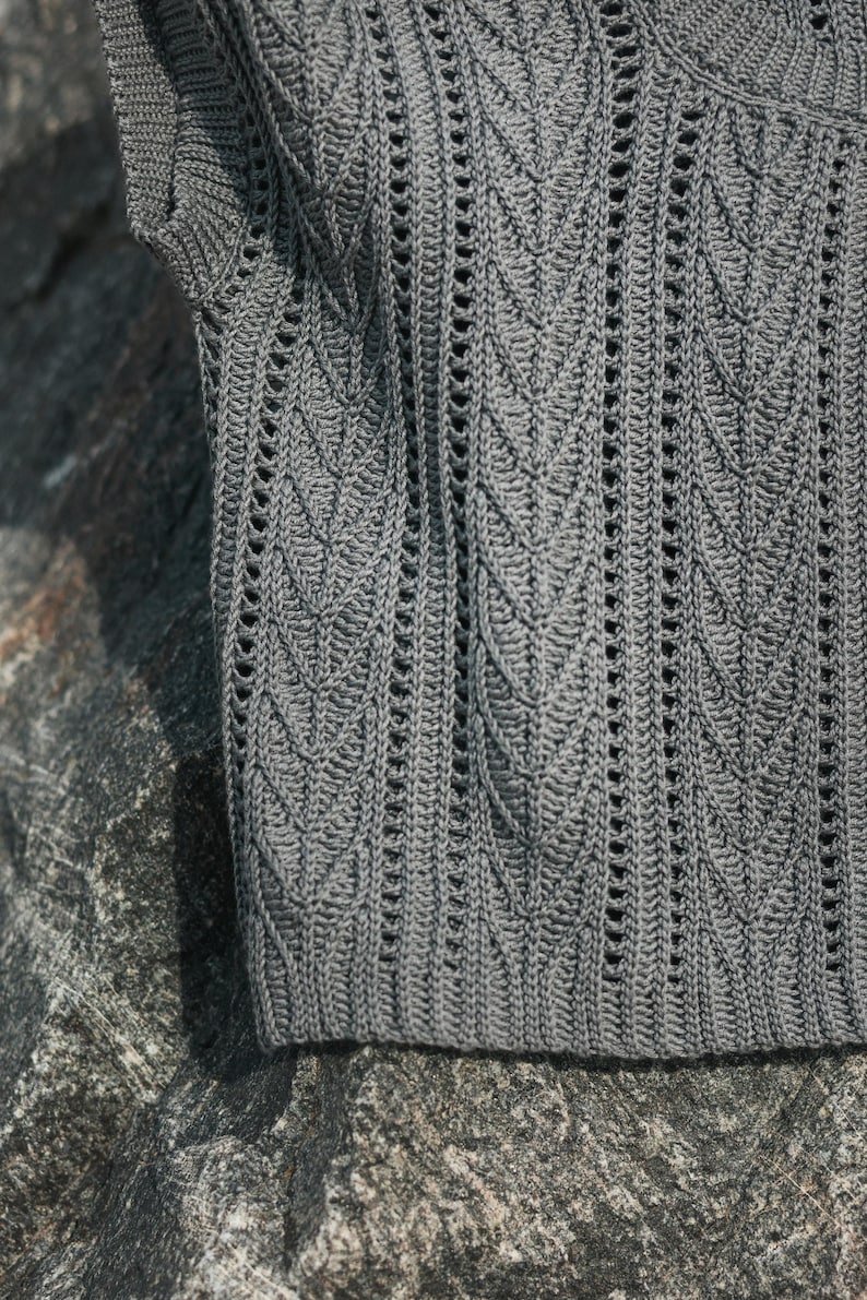 May include: A dark gray sleeveless top featuring a vertical leaf-like crochet pattern with openwork detailing. The garment is constructed with a ribbed hem and a textured knit design that runs throughout the fabric, creating a repeating motif.