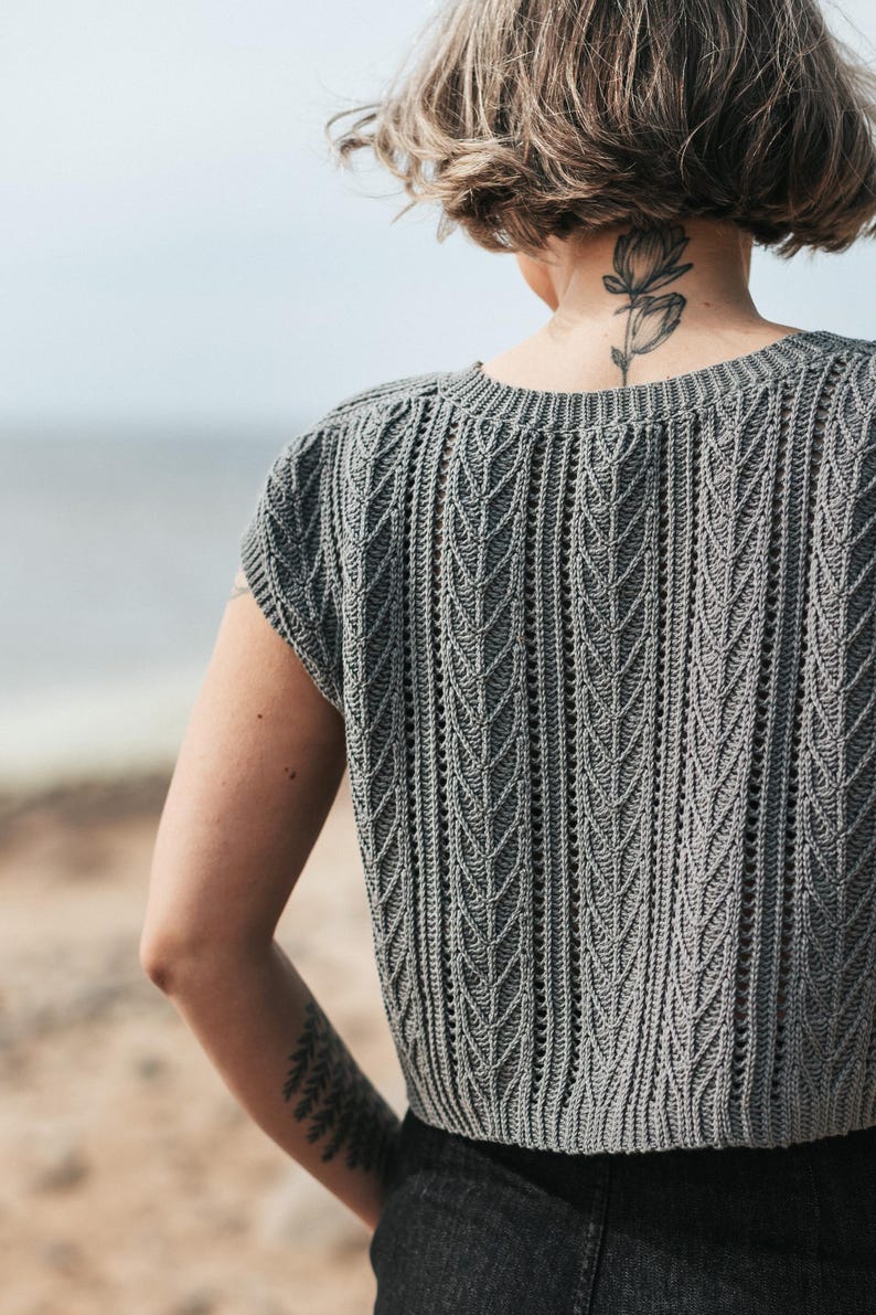 May include: A grey short-sleeved top featuring a vertical knit pattern with repeating leaf-like textures and a round neckline. The garment has a structured ribbed hem and a fitted silhouette, showcasing a detailed stitch design throughout the fabric.