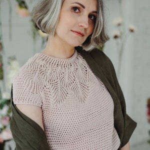May include: A woman with short, gray hair is wearing a light pink crocheted top with a delicate leaf pattern. She is also wearing a dark green linen jacket and matching pants.